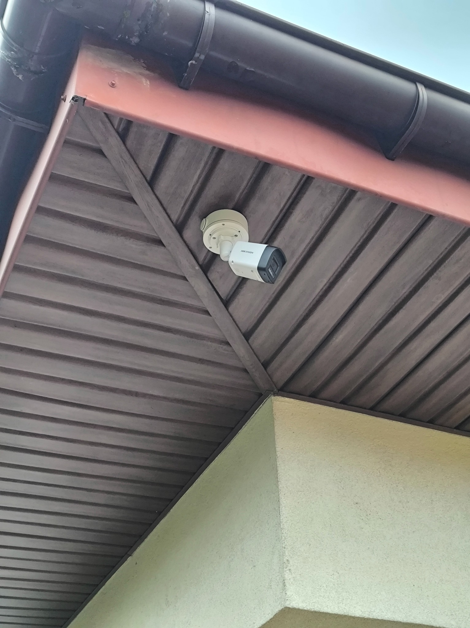 Biała kamera monitoringu Hikvision zamontowana pod brązowym podbitką dachu na zewnątrz budynku, widok z dołu.
