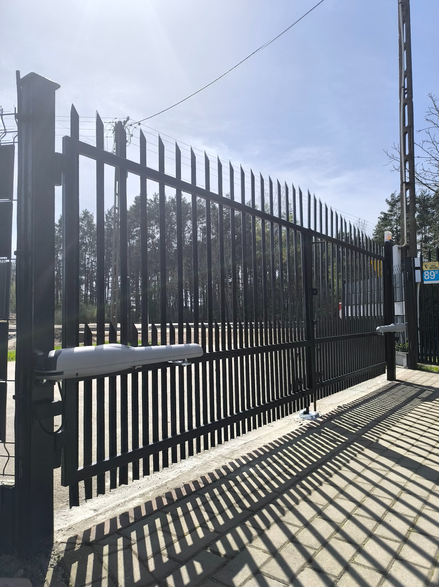 Czarna brama z automatyką, widok z boku, cień rzucany na chodnik, las w tle, słoneczny dzień. Napęd bramy widoczny po lewej stronie.