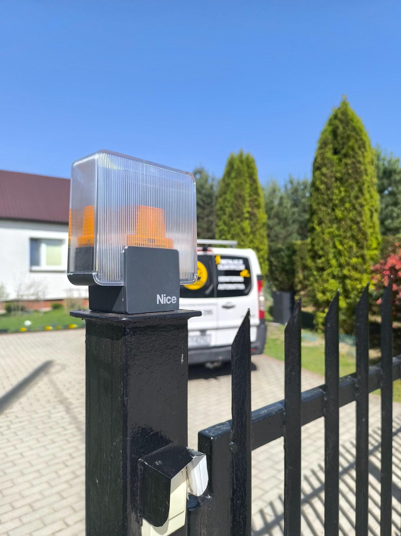 Lampa ostrzegawcza Nice zamontowana na czarnym słupku bramy wjazdowej, z firmowym vanem w tle i zielenią. Słoneczny dzień.