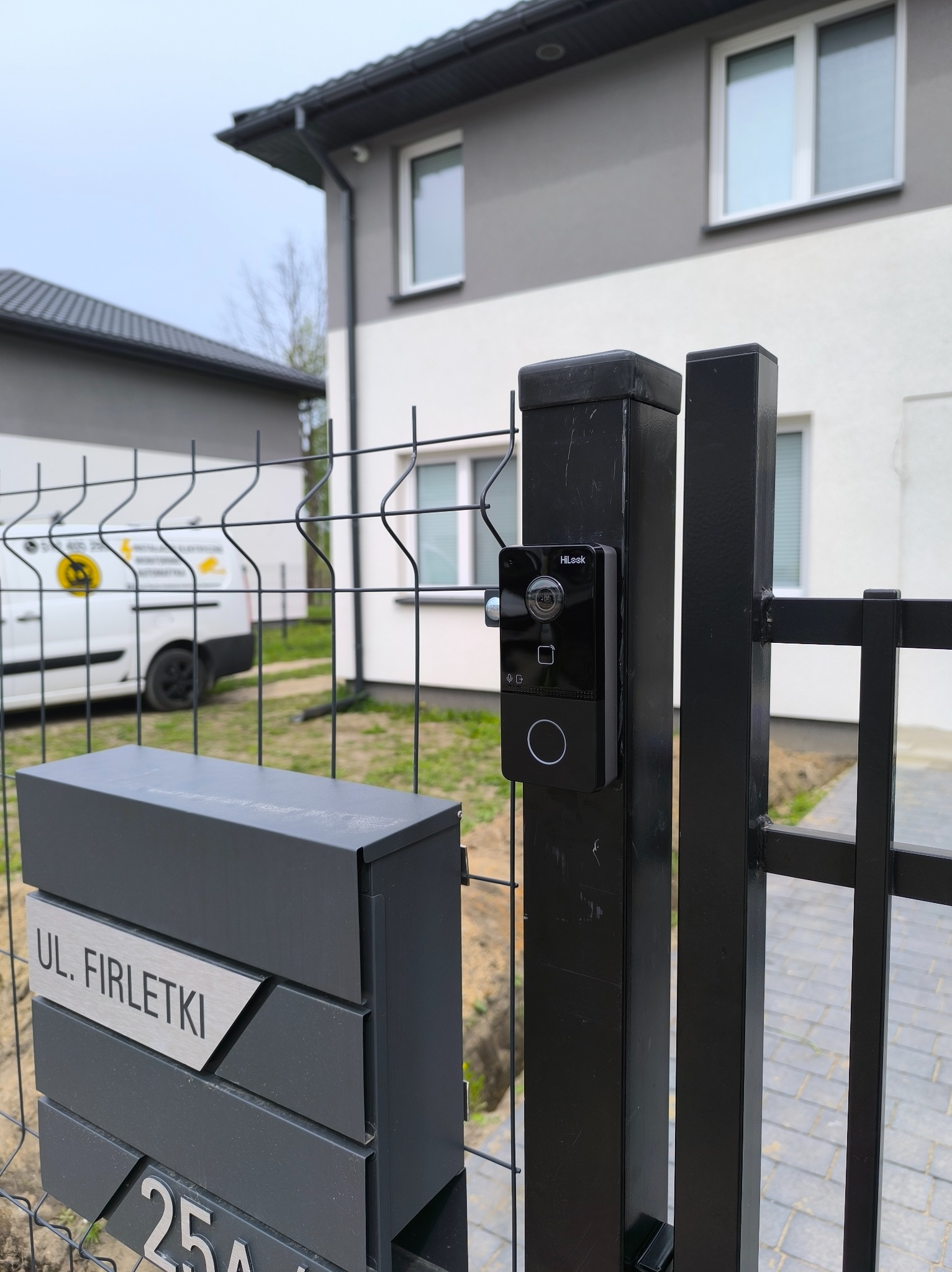 Nowoczesny wideodomofon HiLook zamontowany na czarnym słupku ogrodzeniowym, obok skrzynki pocztowej z adresem, na tle domu i firmowego busa.