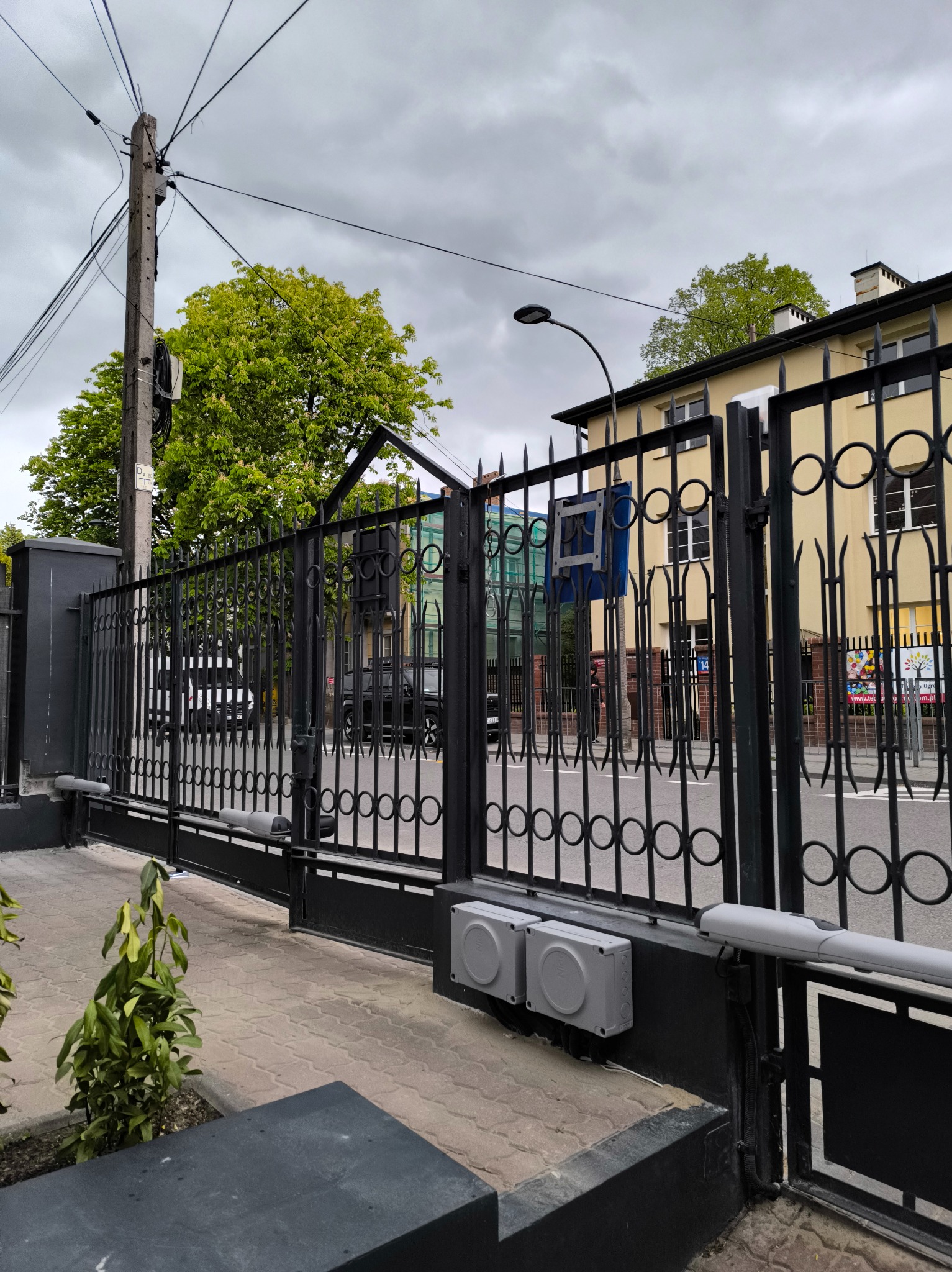 Czarna, metalowa brama wjazdowa z automatyką na tle ulicy i budynku. Widoczne siłowniki i skrzynki sterujące. Pochmurne niebo w tle.