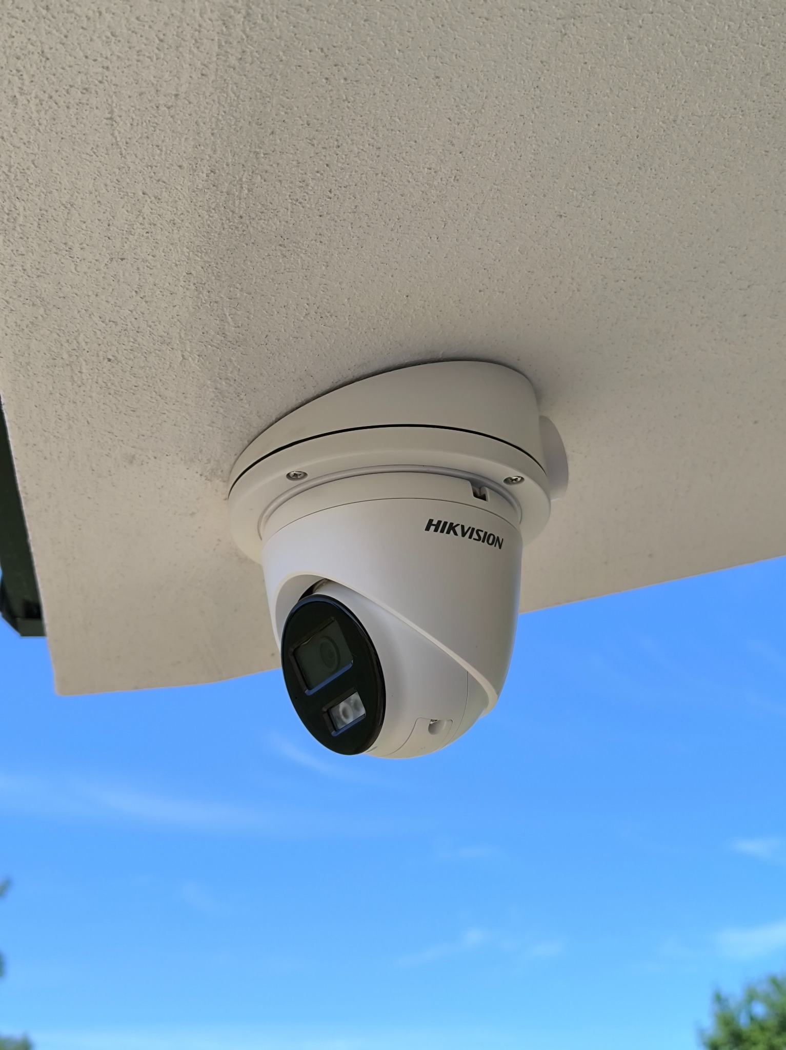 Kamera Hikvision typu turret zamontowana pod jasnym sufitem z widokiem na błękitne niebo. Widoczne mocowanie i logo producenta.