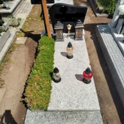 Granitowy nagrobek z równo przyciętym żywopłotem z boku, ozdobiony lampionami i zniczami w różnych kolorach, na cmentarzu.
