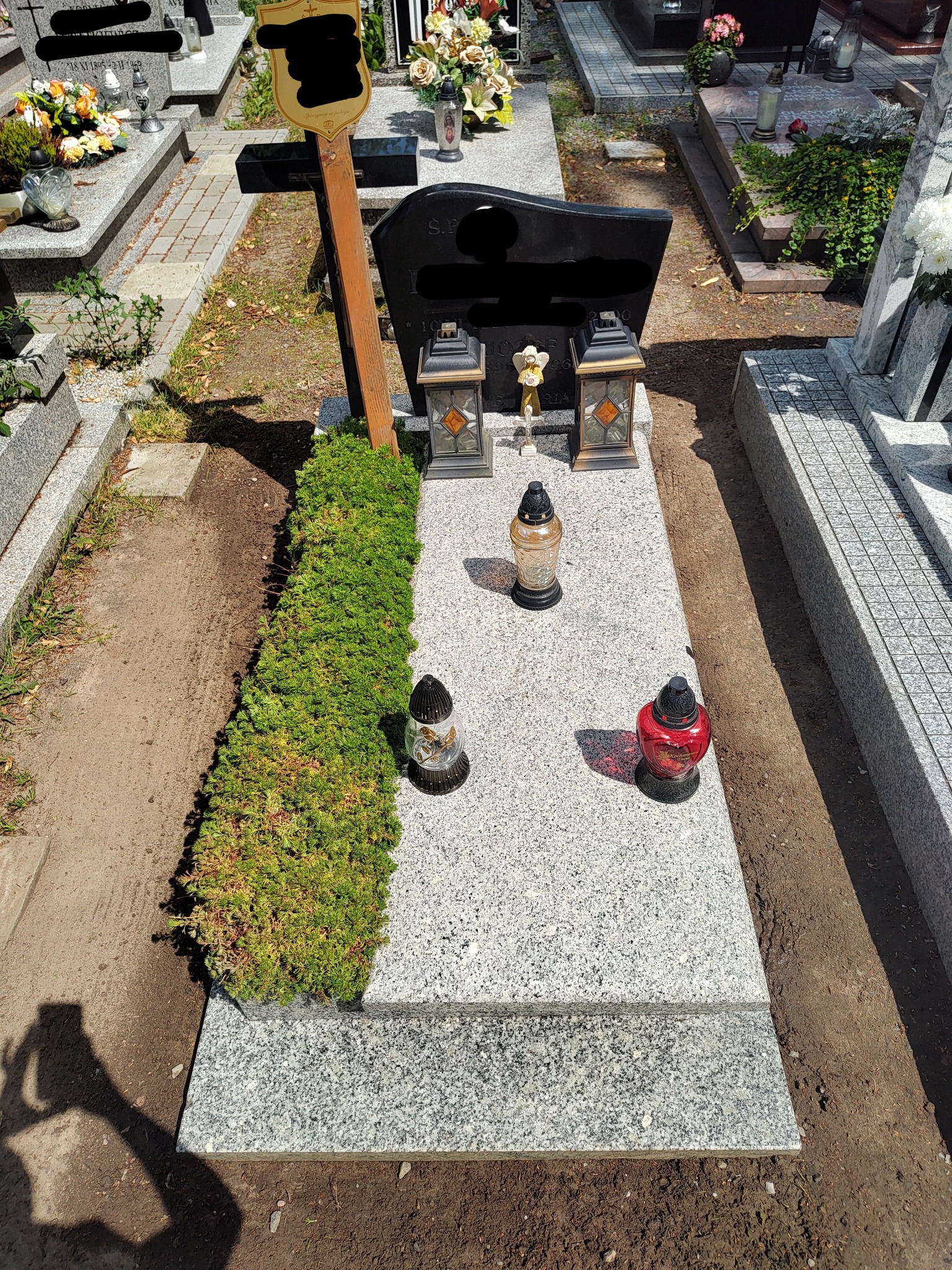 Granitowy nagrobek z równo przyciętym żywopłotem z boku, ozdobiony lampionami i zniczami w różnych kolorach, na cmentarzu.