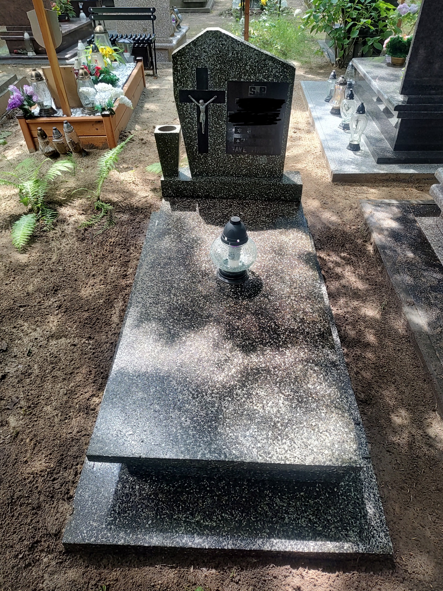 Czarny, granitowy nagrobek z krzyżem i zniczem na płycie, otoczony świeżą ziemią.