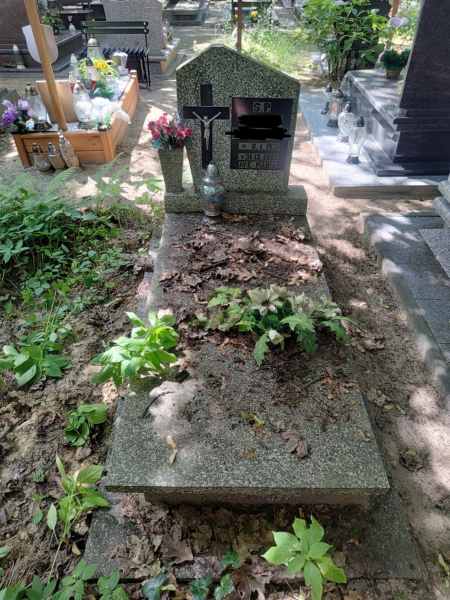 Granitowy nagrobek z krzyżem i wazonem, częściowo pokryty liśćmi i ziemią, otoczony zieloną roślinnością na cmentarzu.