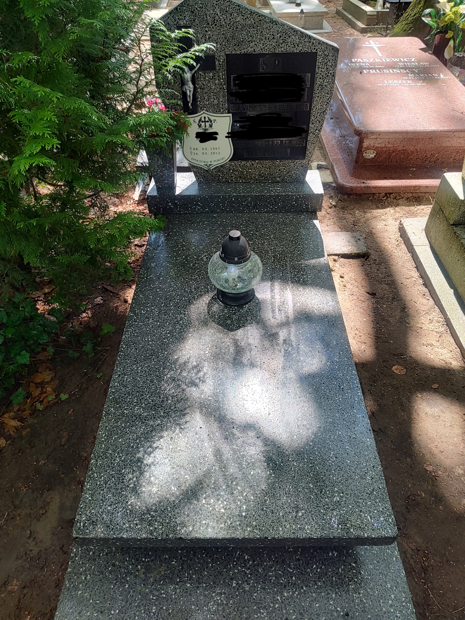 Granitowy nagrobek z lampionem i metalowym krucyfiksem, częściowo ocieniony przez iglaste drzewo, na tle innych nagrobków.