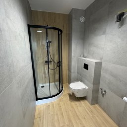 Serhii Diachenko - Nowoczesna łazienka z kabiną prysznicową z czarnymi ramami, wiszącą toaletą i płytkami imitującymi drewno na podłodze oraz szarymi płytkami na ścianach, widoczne zabezpieczenia folią na toalecie...