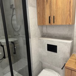 Serhii Diachenko - Nowoczesna łazienka z kabiną prysznicową z czarnymi profilami, wiszącą szafką z drewnianym fornirem, szarą płytką imitującą beton i toaletą w folii ochronnej.