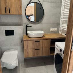 Serhii Diachenko - Wykończona łazienka w szarościach i drewnie: owalne umywalka na blacie, okrągłe lustro z czarnym paskiem, podwieszana szafka z szufladami, toaleta owinięta folią ochronną, pralka z boku.