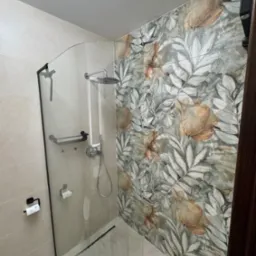 Nowoczesna kabina prysznicowa z czarnymi detalami i szklaną ścianką, wyłożona płytkami w kwiatowy wzór. Widoczny odpływ liniowy i uchwyt na papier toaletowy.