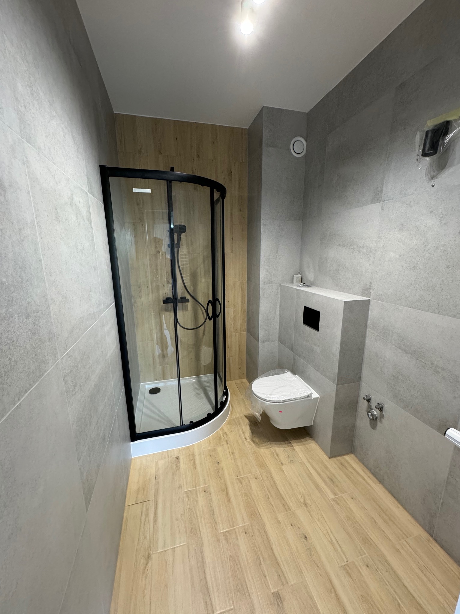 Nowoczesna łazienka z kabiną prysznicową z czarnymi ramami, wiszącą toaletą i płytkami imitującymi drewno na podłodze oraz szarymi płytkami na ścianach, widoczne zabezpieczenia folią na toalecie...