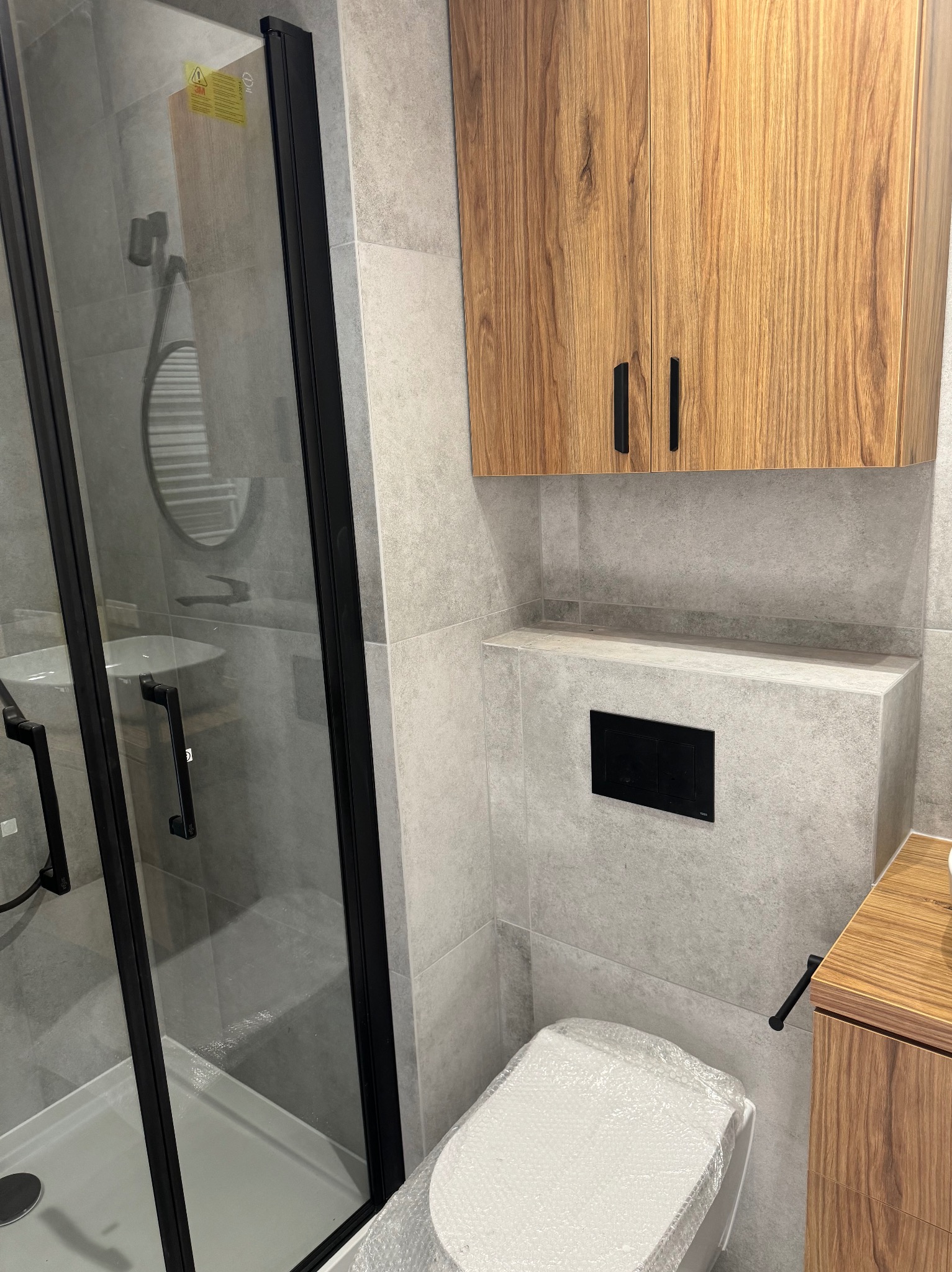 Nowoczesna łazienka z kabiną prysznicową z czarnymi profilami, wiszącą szafką z drewnianym fornirem, szarą płytką imitującą beton i toaletą w folii ochronnej.