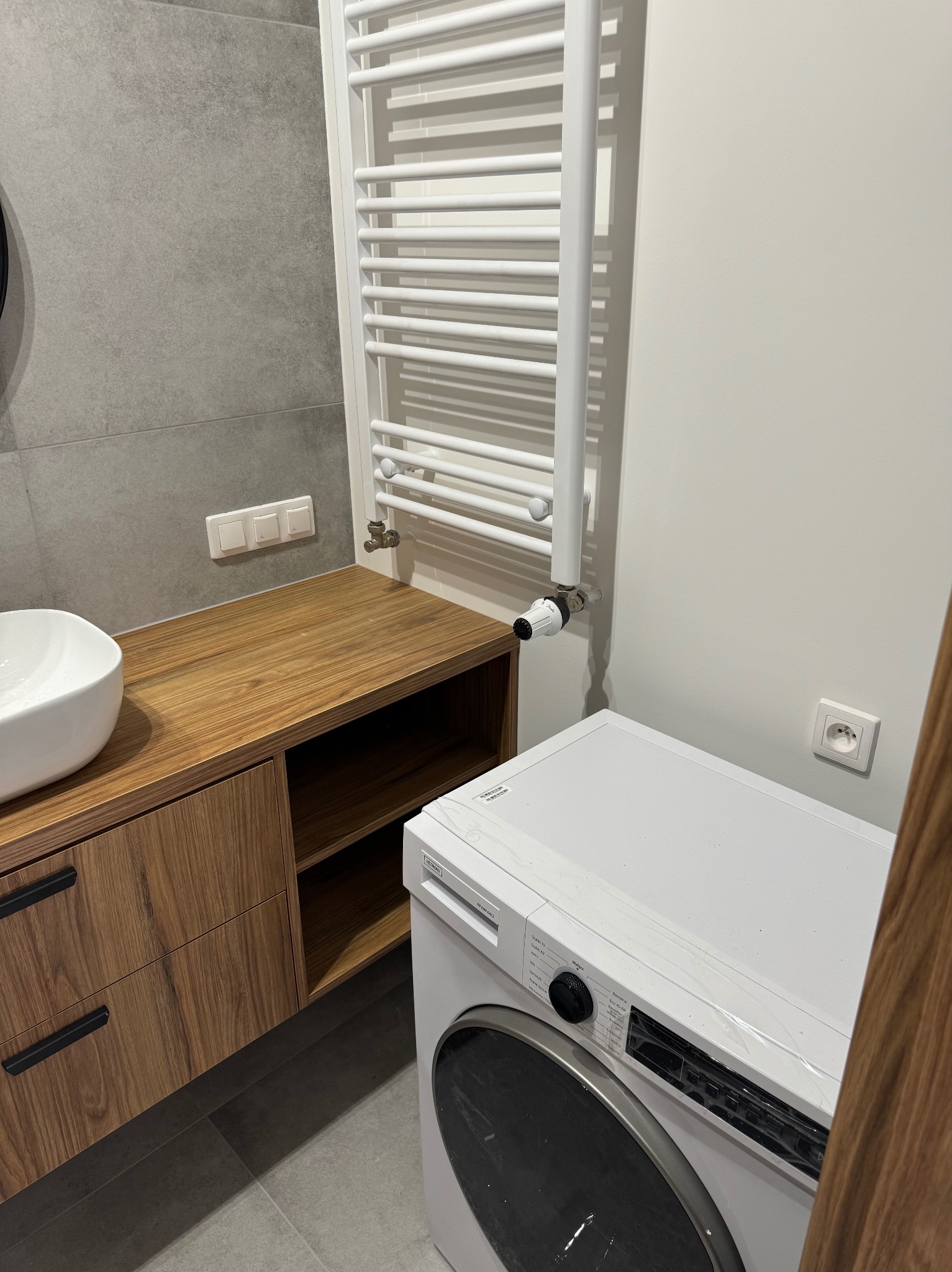 Nowoczesna łazienka z drewnianą szafką pod umywalką, grzejnikiem drabinkowym i białą pralką.