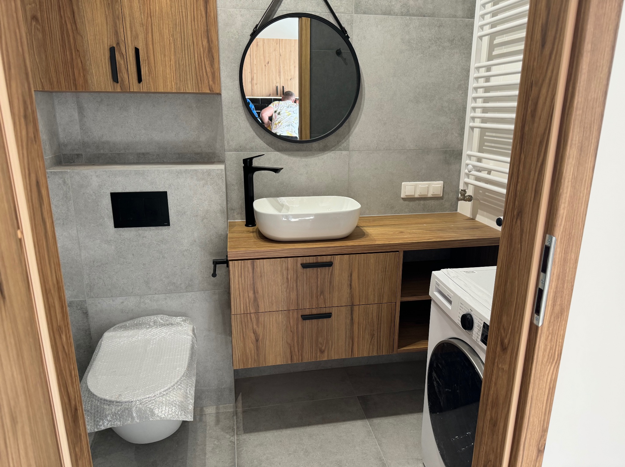 Wykończona łazienka w szarościach i drewnie: owalne umywalka na blacie, okrągłe lustro z czarnym paskiem, podwieszana szafka z szufladami, toaleta owinięta folią ochronną, pralka z boku.