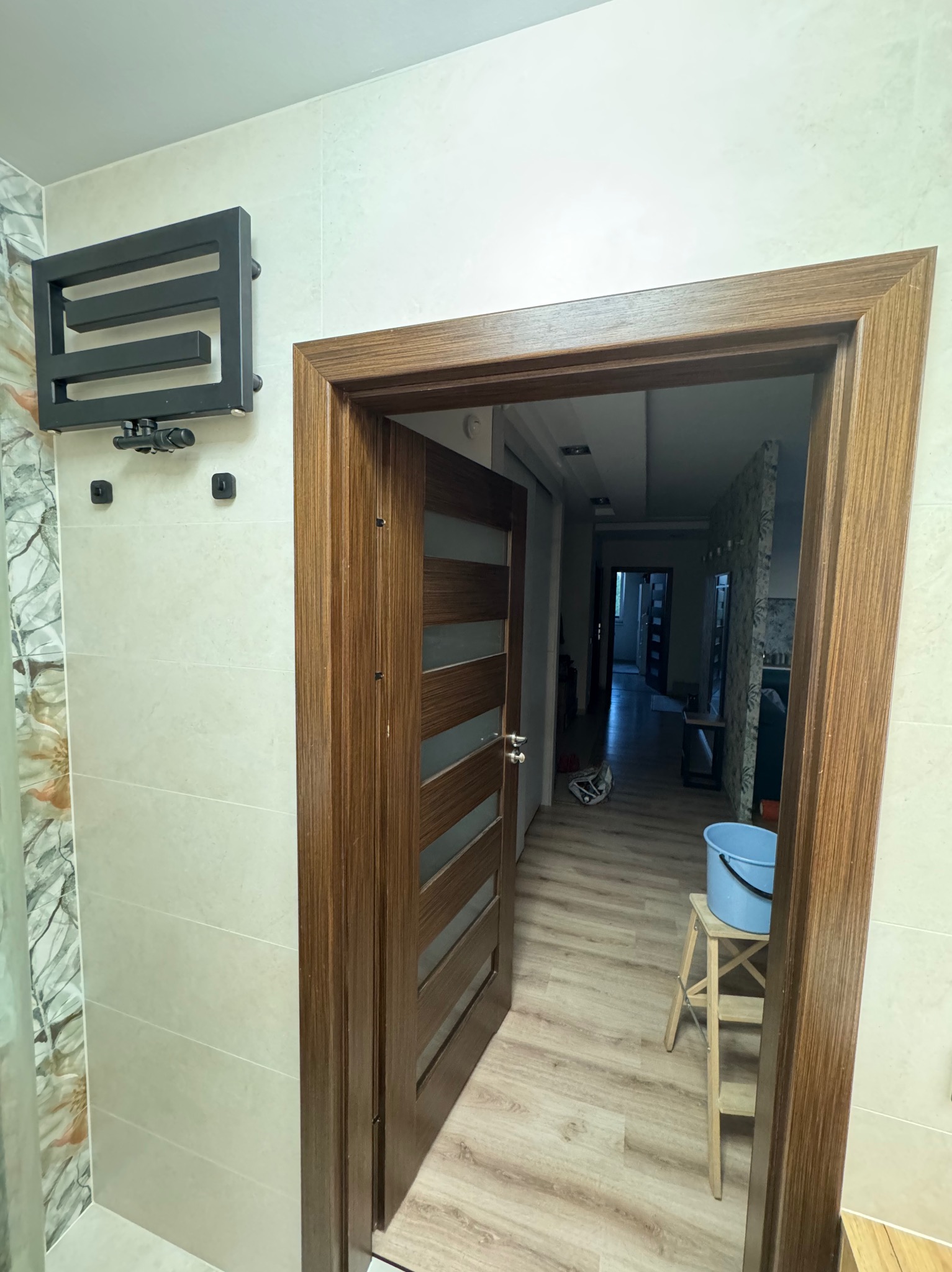 Otwarta, drewniana, nowoczesna rama drzwiowa z przeszkleniami w łazience z grzejnikiem drabinkowym i beżowymi płytkami. Widok na dalsze pomieszczenia.