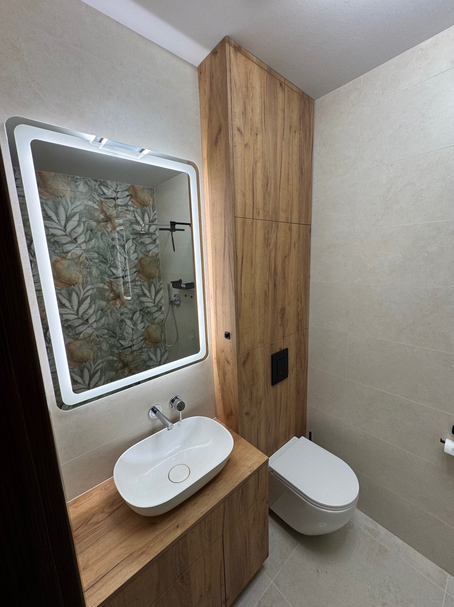 Nowoczesna łazienka z drewnianą szafką pod umywalką, podświetlanym lustrem i wiszącą toaletą. Widoczna armatura w kolorze chromu i płytki w odcieniach beżu.