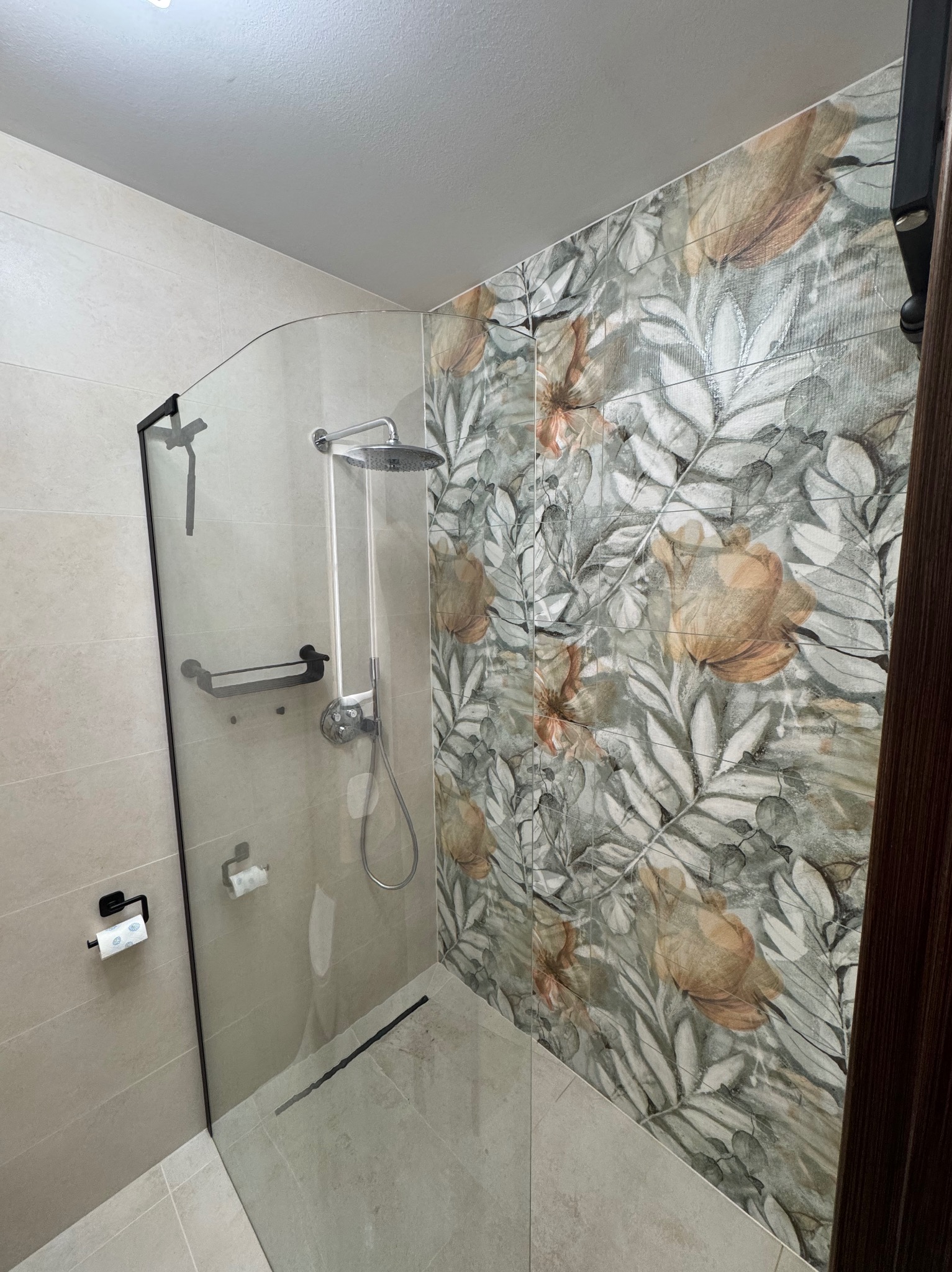 Nowoczesna kabina prysznicowa z czarnymi detalami i szklaną ścianką, wyłożona płytkami w kwiatowy wzór. Widoczny odpływ liniowy i uchwyt na papier toaletowy.