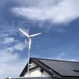 Mała turbina wiatrowa z zielonymi końcówkami łopat zamontowana na dachu budynku obok paneli słonecznych, na tle błękitnego nieba z chmurami.