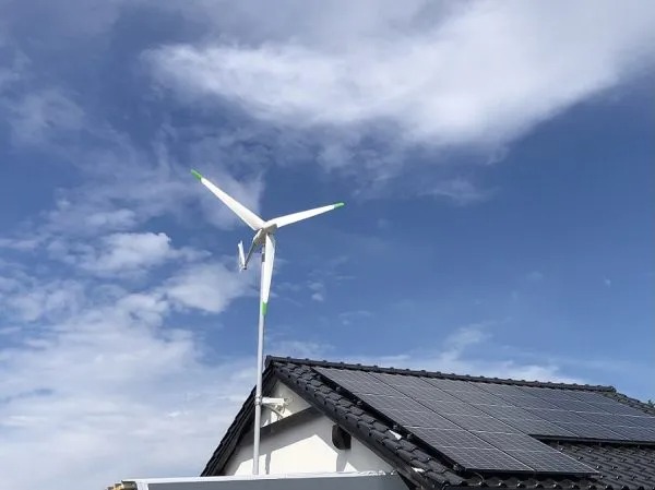 Mała turbina wiatrowa z zielonymi końcówkami łopat zamontowana na dachu budynku obok paneli słonecznych, na tle błękitnego nieba z chmurami.