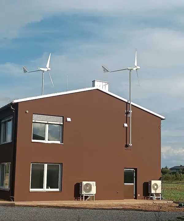 Dwa białe generatory wiatrowe zamontowane na dachu dwukondygnacyjnego domu o brązowej elewacji, z dwoma zewnętrznymi jednostkami klimatyzacji przy ścianie i oknami z żaluzjami.