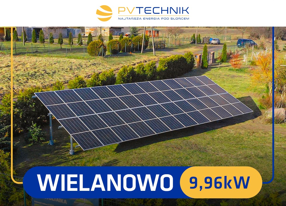 Instalacja paneli słonecznych na trawiastym terenie w Wielanowie, z widocznymi budynkami i drzewami w tle oraz dwoma zaparkowanymi samochodami.