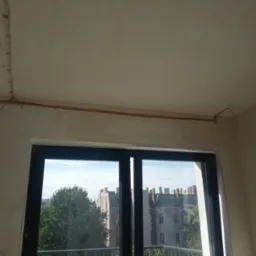 Czerwony przewód elektryczny poprowadzony wzdłuż sufitu i ściany, widoczny w trakcie prac instalacyjnych w pomieszczeniu z oknem balkonowym, w tle budynki.