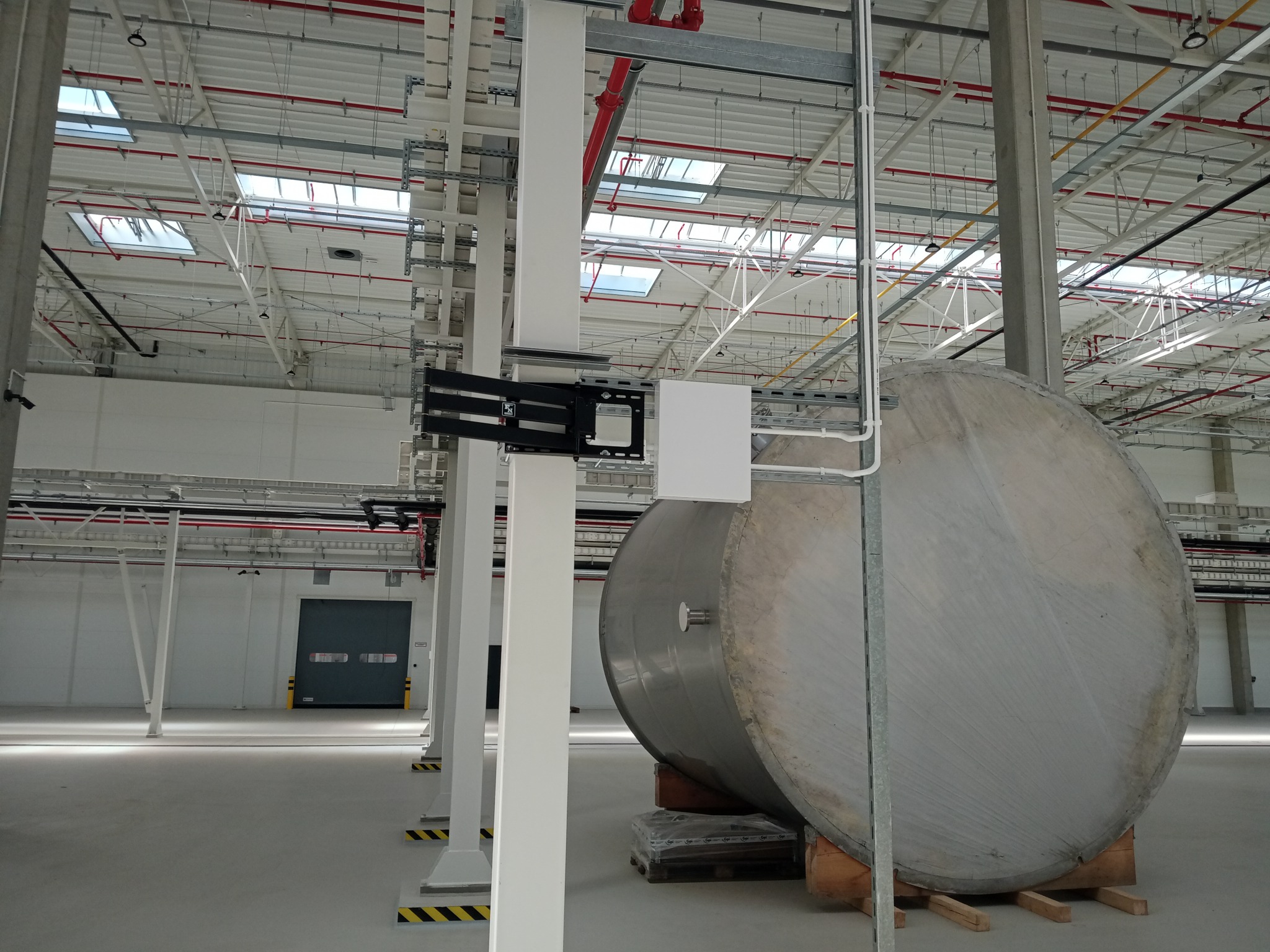 Wnętrze hali przemysłowej z widocznymi instalacjami pod sufitem, rurami, kablami oraz dużym, cylindrycznym zbiornikiem na paletach, z zamontowaną skrzynką elektryczną na słupie.