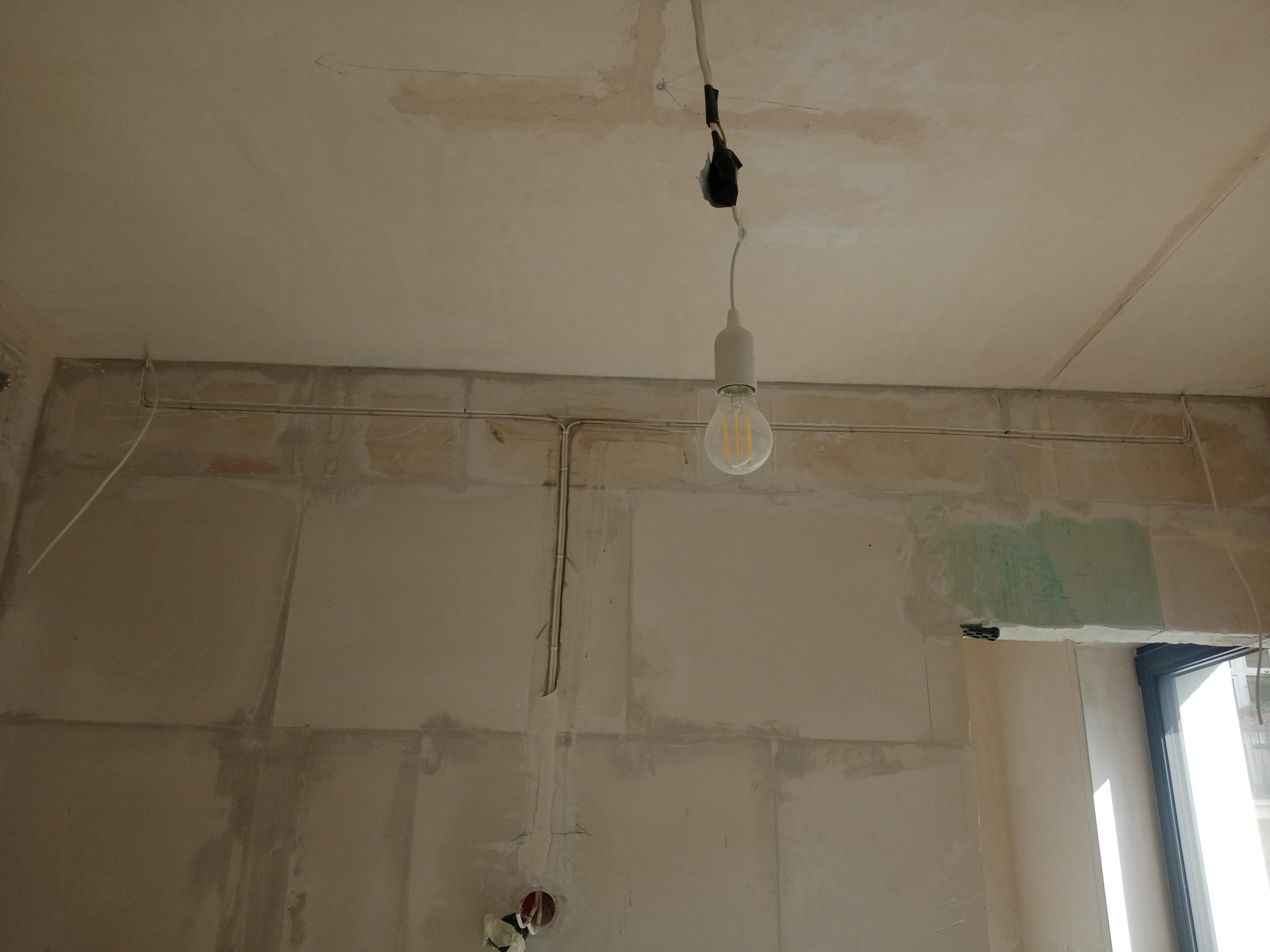 Niedokończone wnętrze z widocznymi przewodami elektrycznymi na ścianie, surowy tynk, wisząca żarówka z prowizorycznym podłączeniem, widoczne puszki elektryczne.