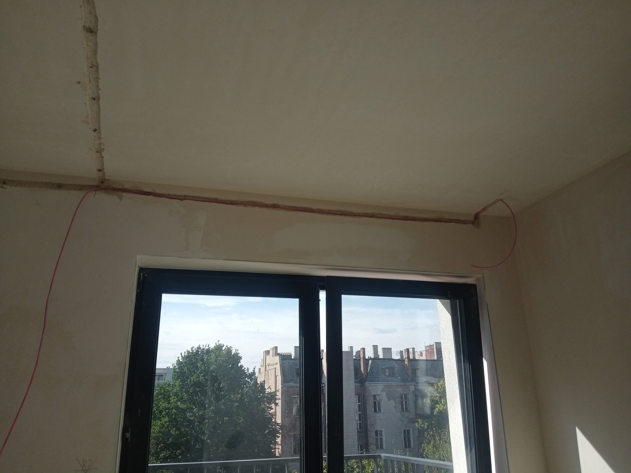 Czerwony przewód elektryczny poprowadzony wzdłuż sufitu i ściany, widoczny w trakcie prac instalacyjnych w pomieszczeniu z oknem balkonowym, w tle budynki.