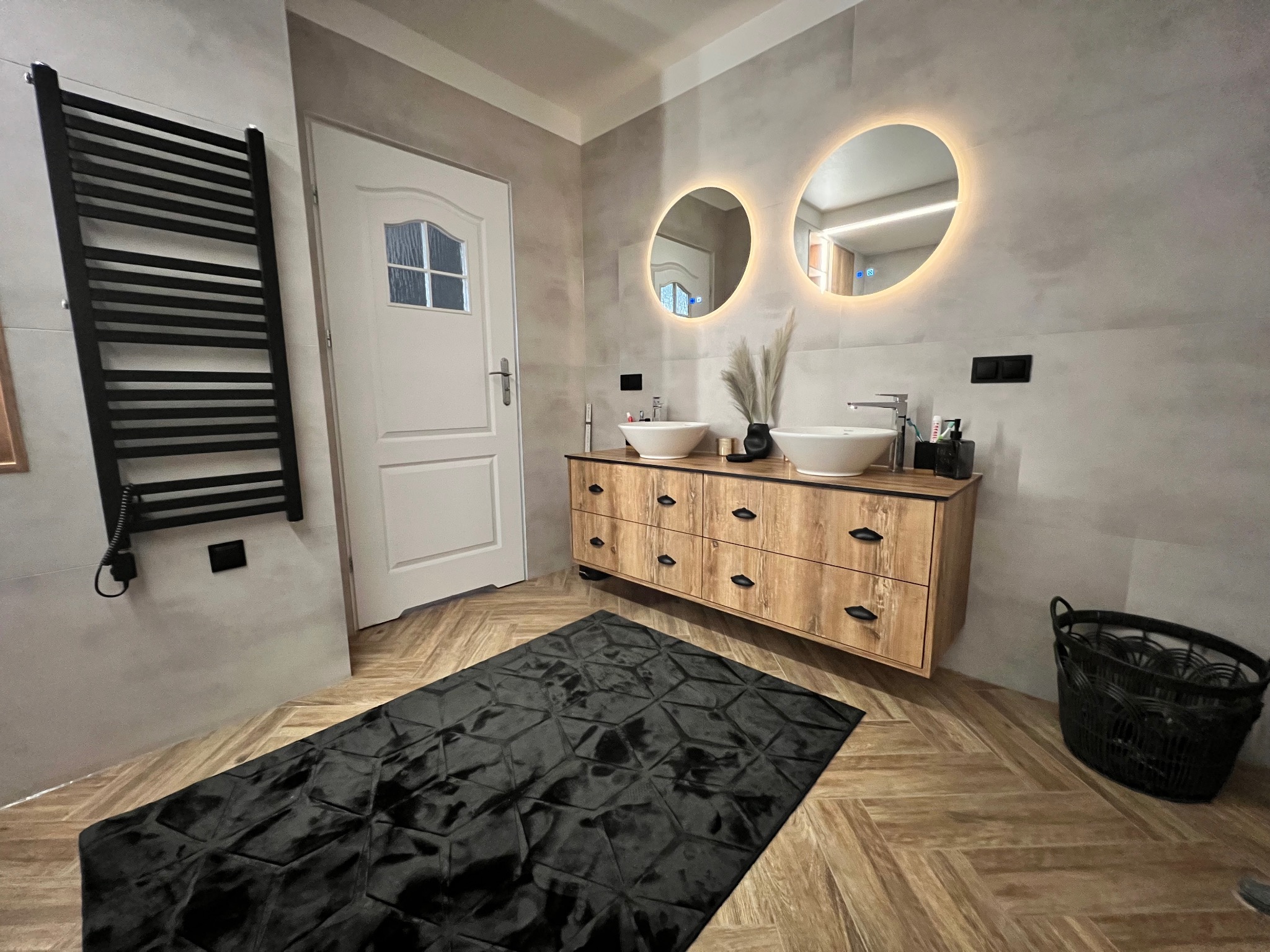 Nowoczesna łazienka z dwoma okrągłymi lustrami podświetlanymi LED, drewnianą szafką pod umywalki nablatowe, czarnym grzejnikiem drabinkowym i wzorzystym dywanem na podłodze w jodełkę.