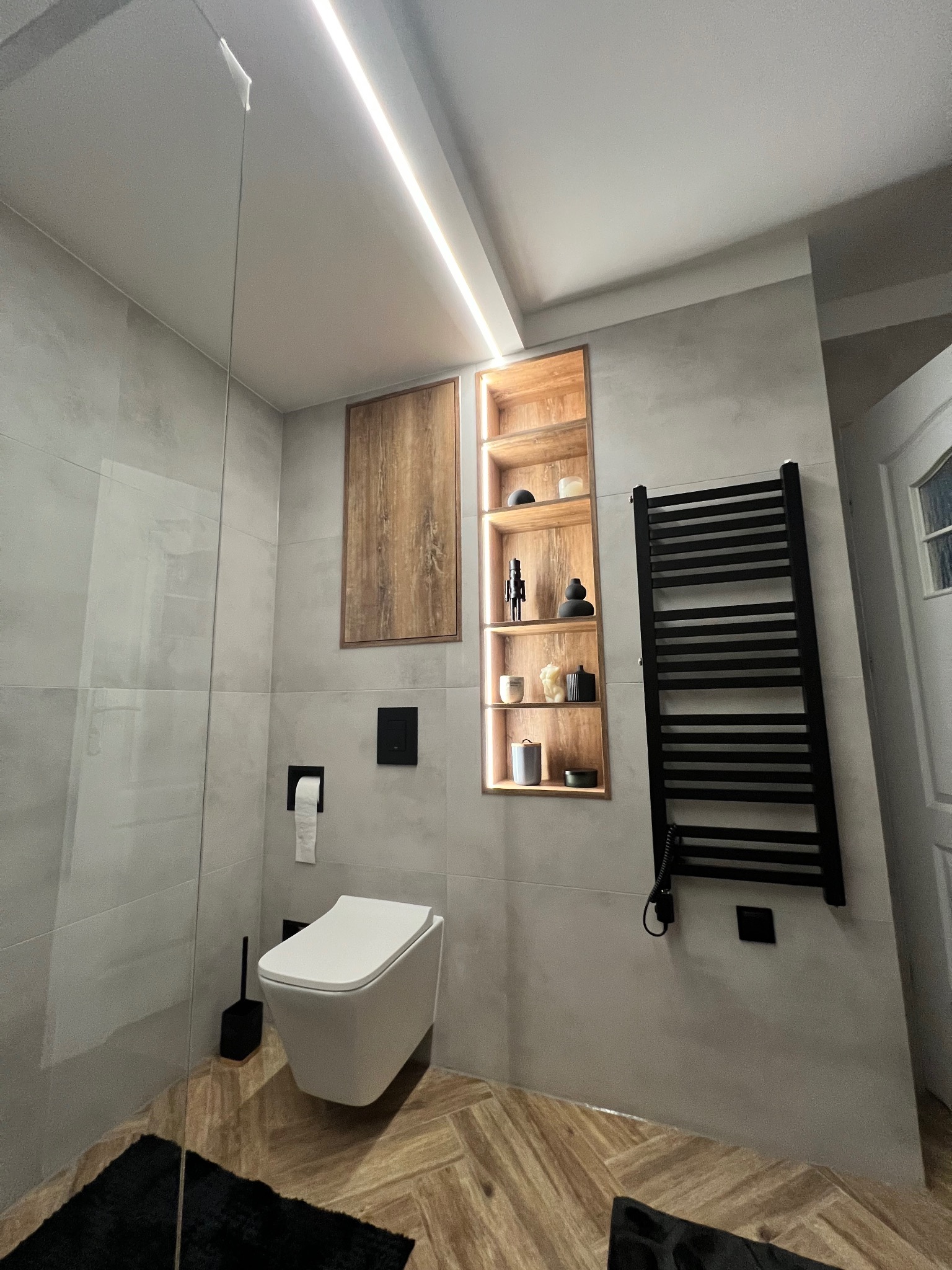 Nowoczesna łazienka z szarymi płytkami, podświetlaną wnęką z dekoracjami, wiszącą toaletą i czarnym grzejnikiem drabinkowym.