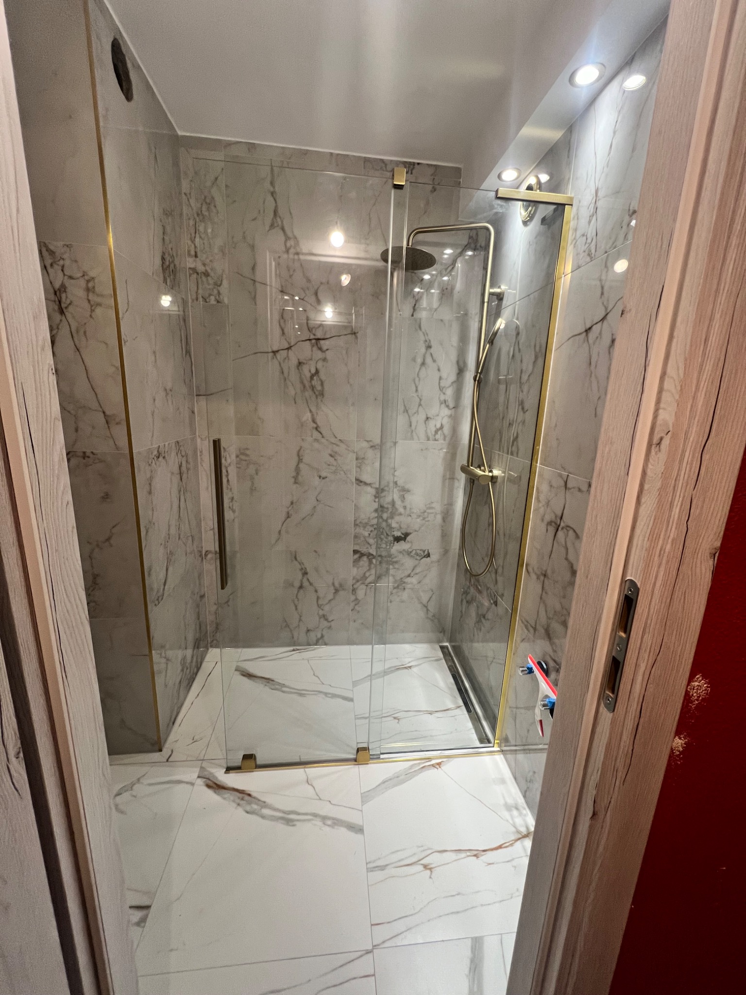 Nowoczesna kabina prysznicowa z marmurowymi płytkami w odcieniach szarości i białej podłodze z marmurowym wzorem, złote wykończenia, widok zza otwartych drewnianych drzwi.