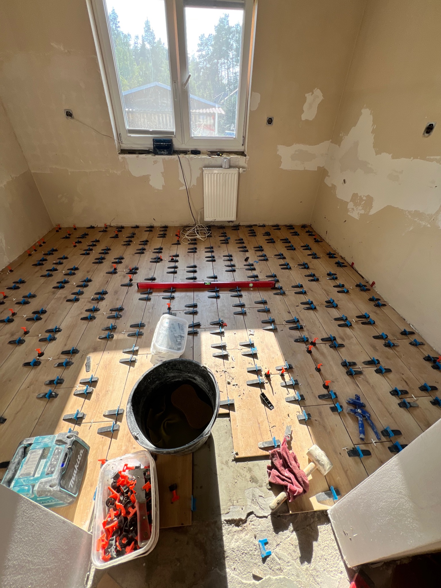 Podłoga w trakcie układania płytek, widoczne krzyżyki dystansowe, poziomnica, wiadro z zaprawą, gumowy młotek i narzędzia, ściany przygotowane pod malowanie, okno dachowe.