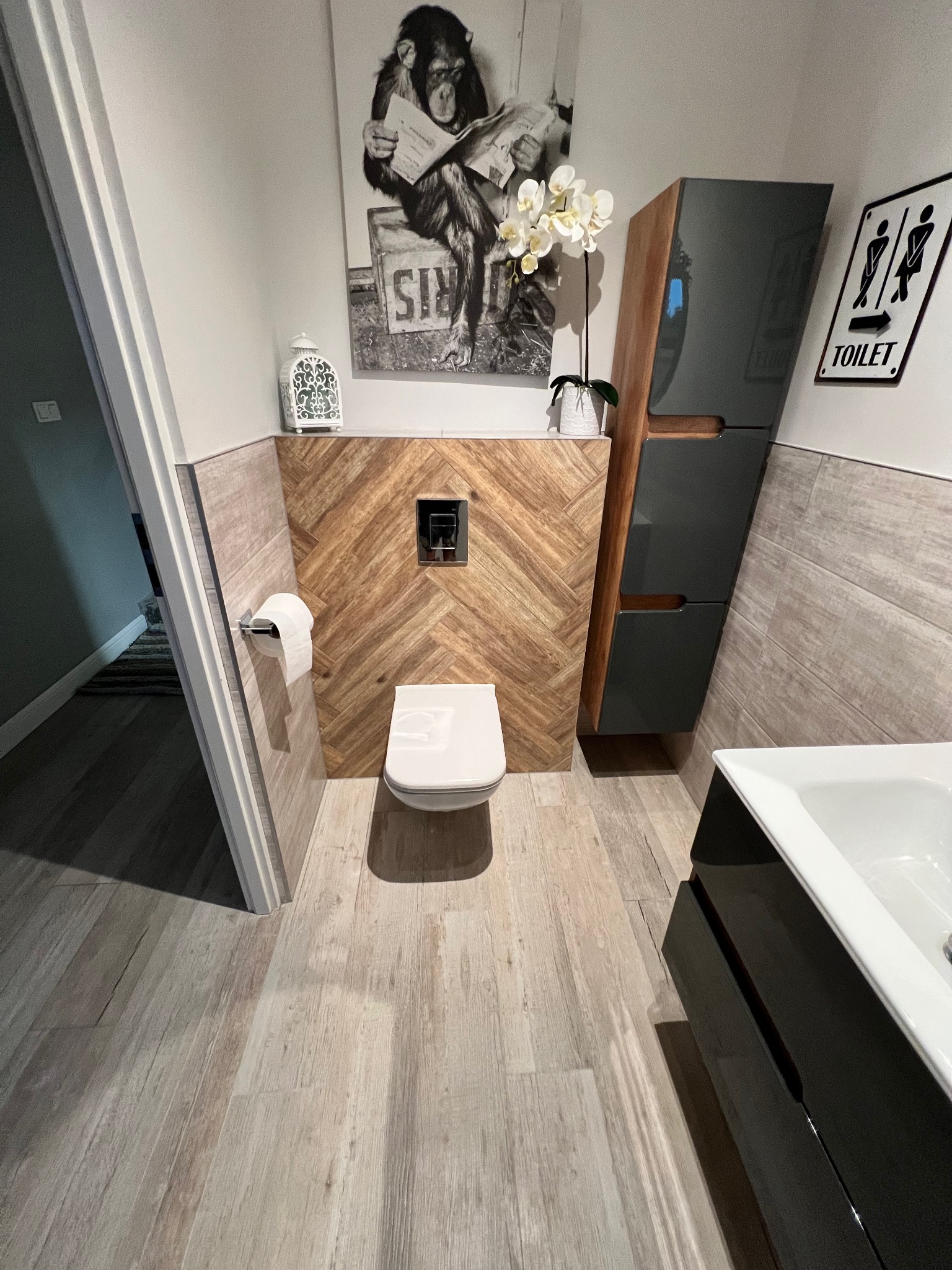 Nowoczesna łazienka z drewnopodobnymi płytkami ułożonymi w jodełkę na ścianie za toaletą, wisząca toaleta, szara szafka łazienkowa, umywalka z czarnym blatem, plakat z szympansem czytającym gazetę...