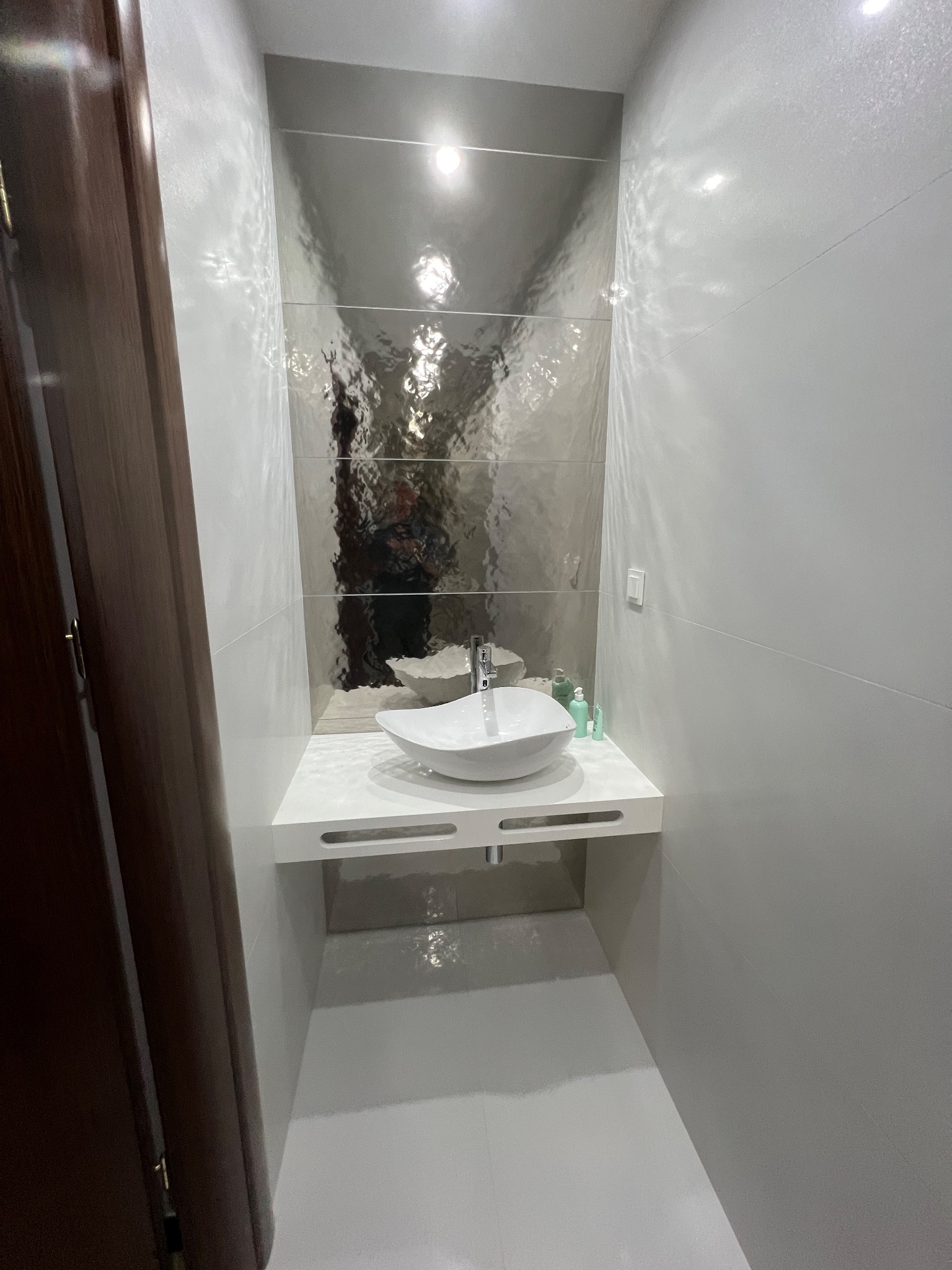 Nowoczesna łazienka w odcieniach szarości z oryginalną umywalką i błyszczącymi płytkami na ścianie. Widoczne akcesoria łazienkowe i ciemne drzwi po lewej.