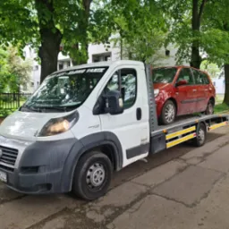 Biała laweta z czerwonym uszkodzonym samochodem osobowym na platformie, zaparkowana na ulicy w słoneczny dzień, widoczne drzewa i budynek w tle.