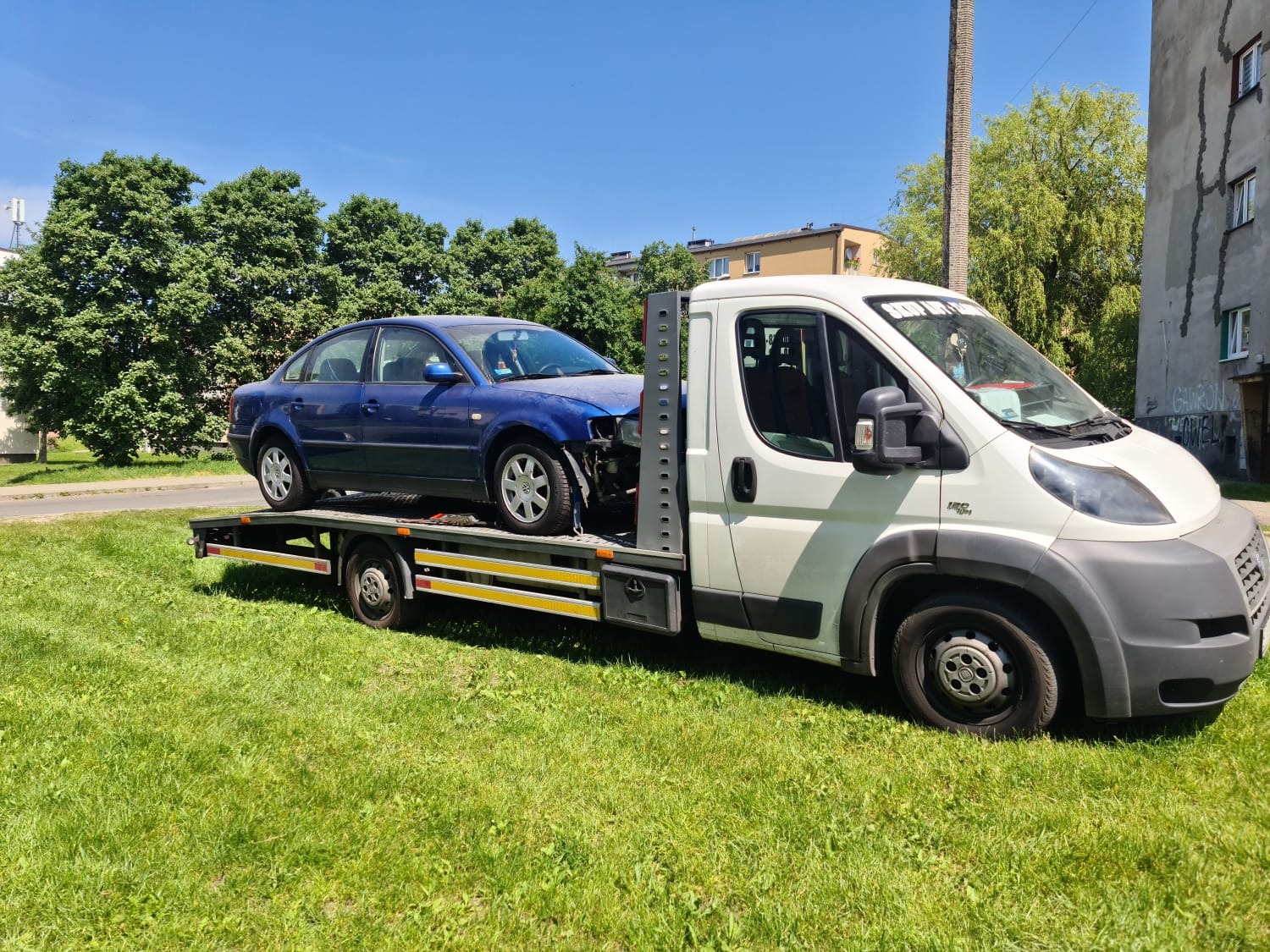 Niebieski samochód osobowy z uszkodzonym przodem, transportowany na lawecie na tle zielonego trawnika i budynków mieszkalnych w słoneczny dzień.