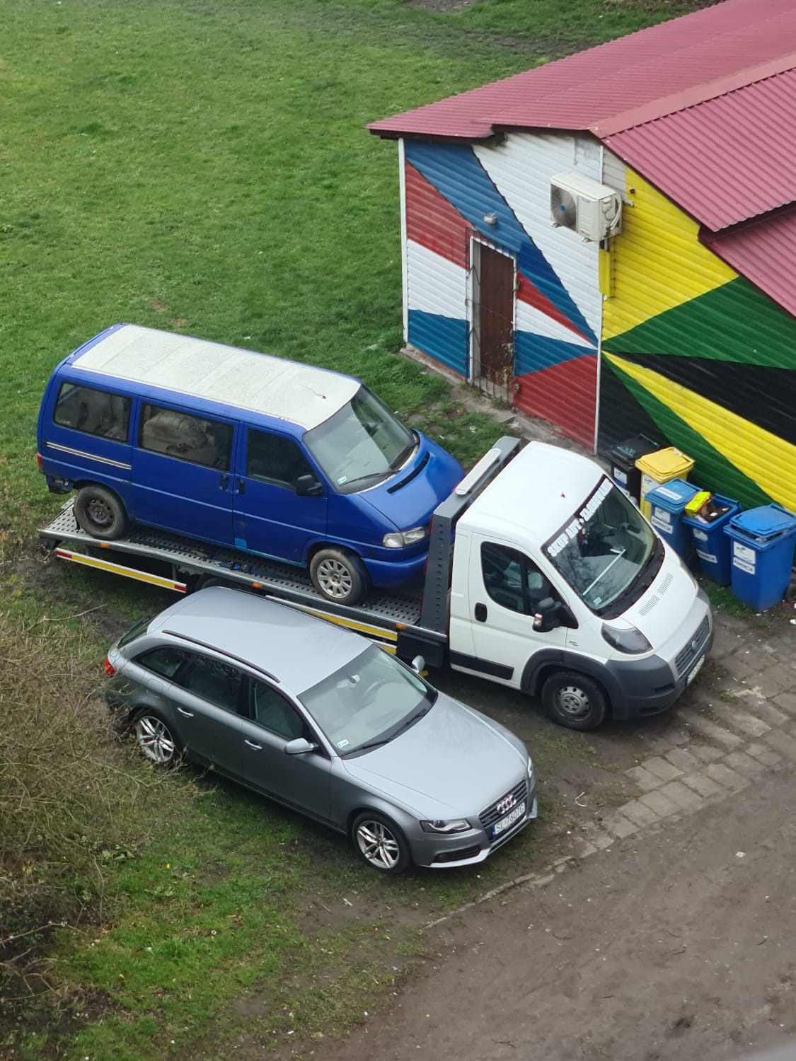 Dwa samochody - szary sedan i niebieski van - umieszczone na platformie białej lawety na tle zielonego trawnika i budynku z pomalowanymi ścianami w barwach flag.