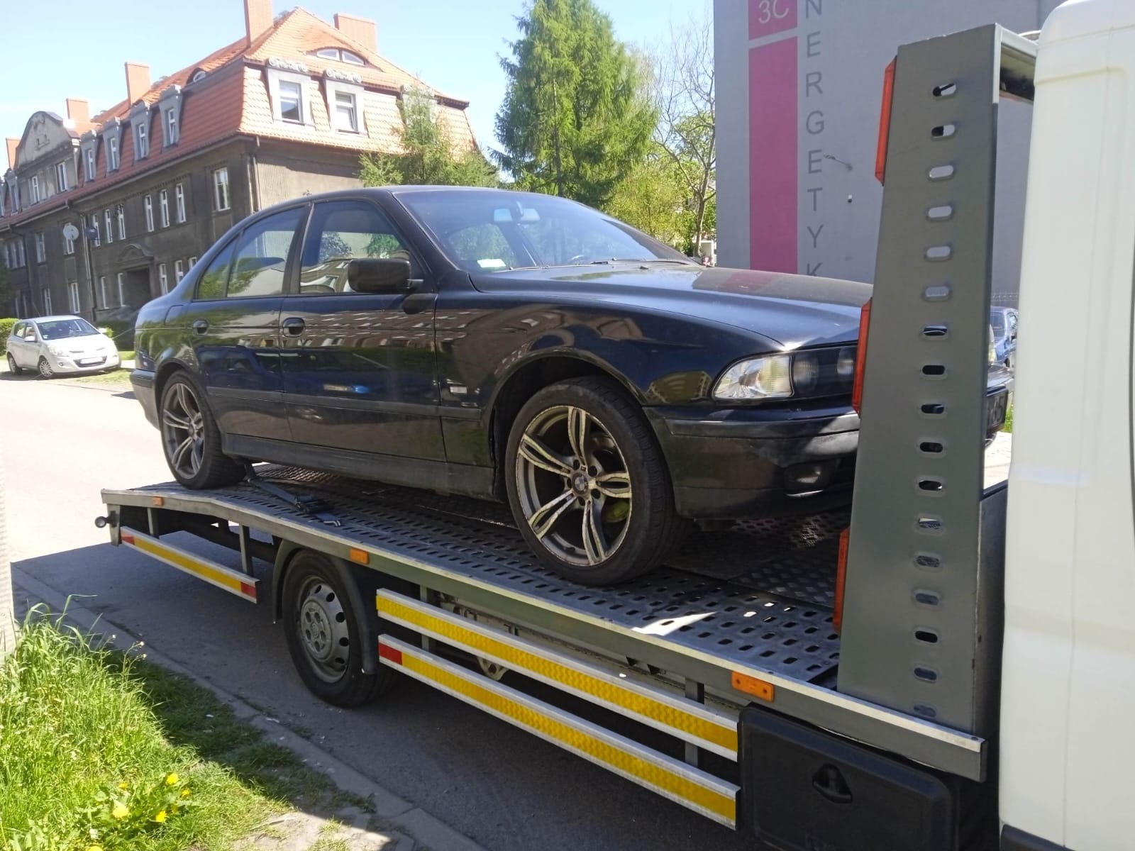 Czarny samochód osobowy marki BMW E39 na platformie lawety, z widocznymi elementami mocującymi i żółtymi pasami odblaskowymi, w tle budynek mieszkalny.