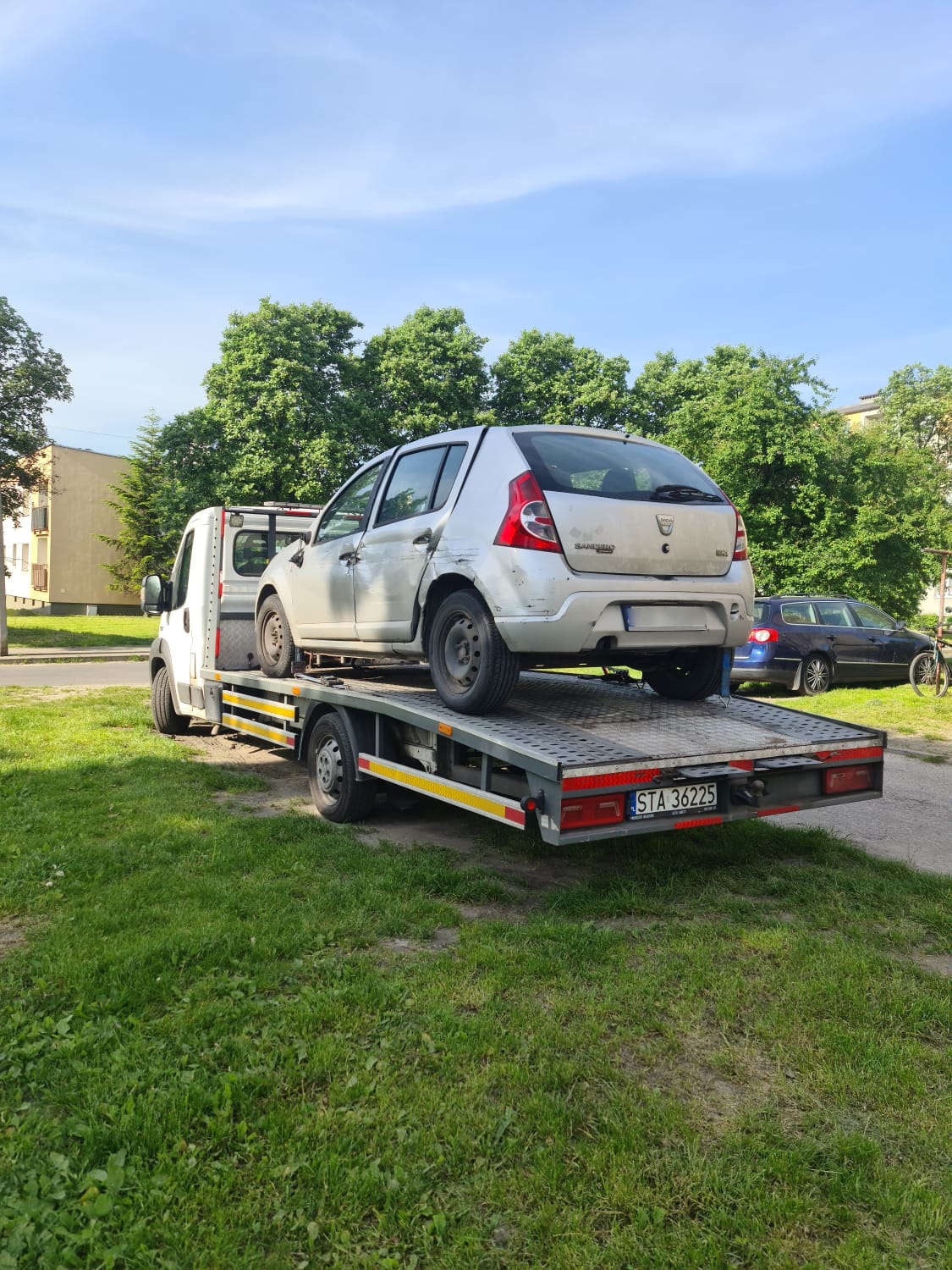 Uszkodzony srebrny Dacia Sandero na platformie lawety, widok z tyłu, na tle trawnika i budynków.
