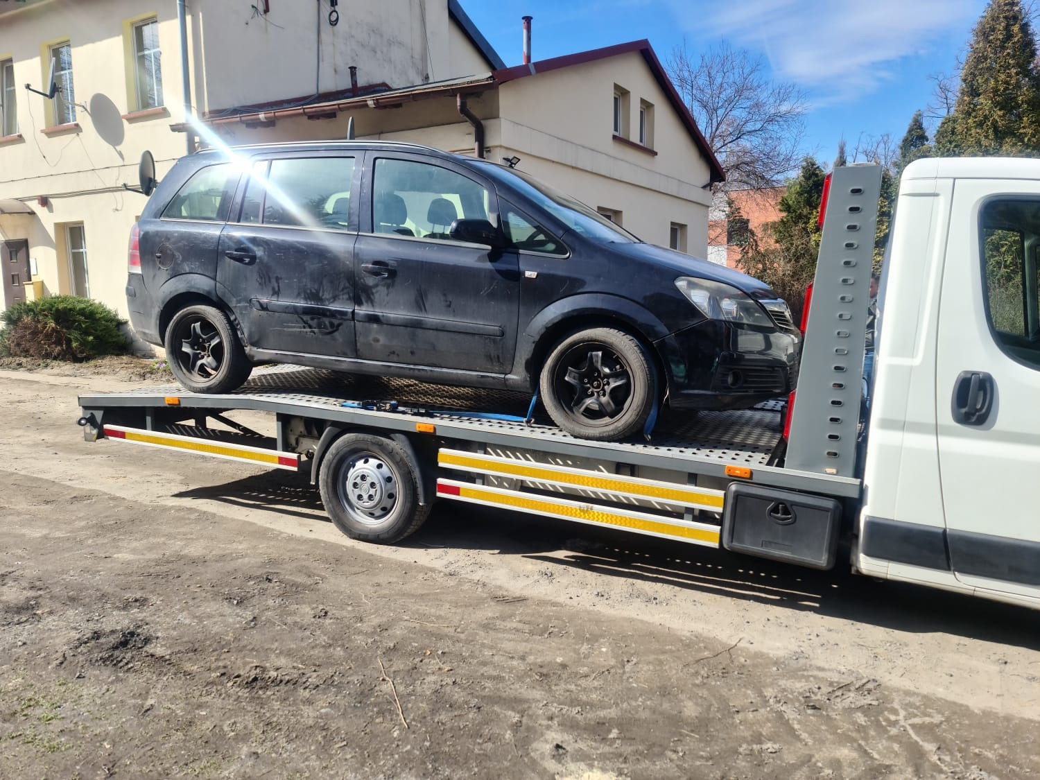 Czarny, lekko uszkodzony samochód osobowy typu kombi, zabezpieczony pasami transportowymi, przewożony na platformie lawety w słoneczny dzień. W tle widoczny budynek mieszkalny.
