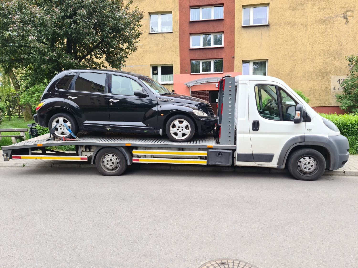 Czarny Chrysler PT Cruiser zabezpieczony pasami transportowymi na platformie białej lawety, na tle bloku mieszkalnego z numerem 42.