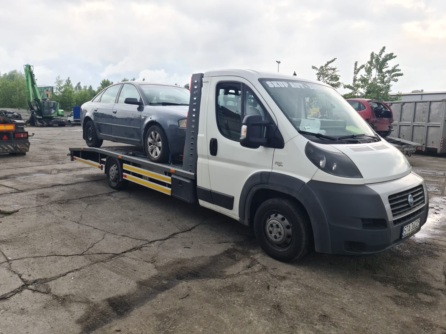 Szary samochód osobowy przewożony na białej lawecie na betonowym placu z widocznym złomowiskiem w tle.