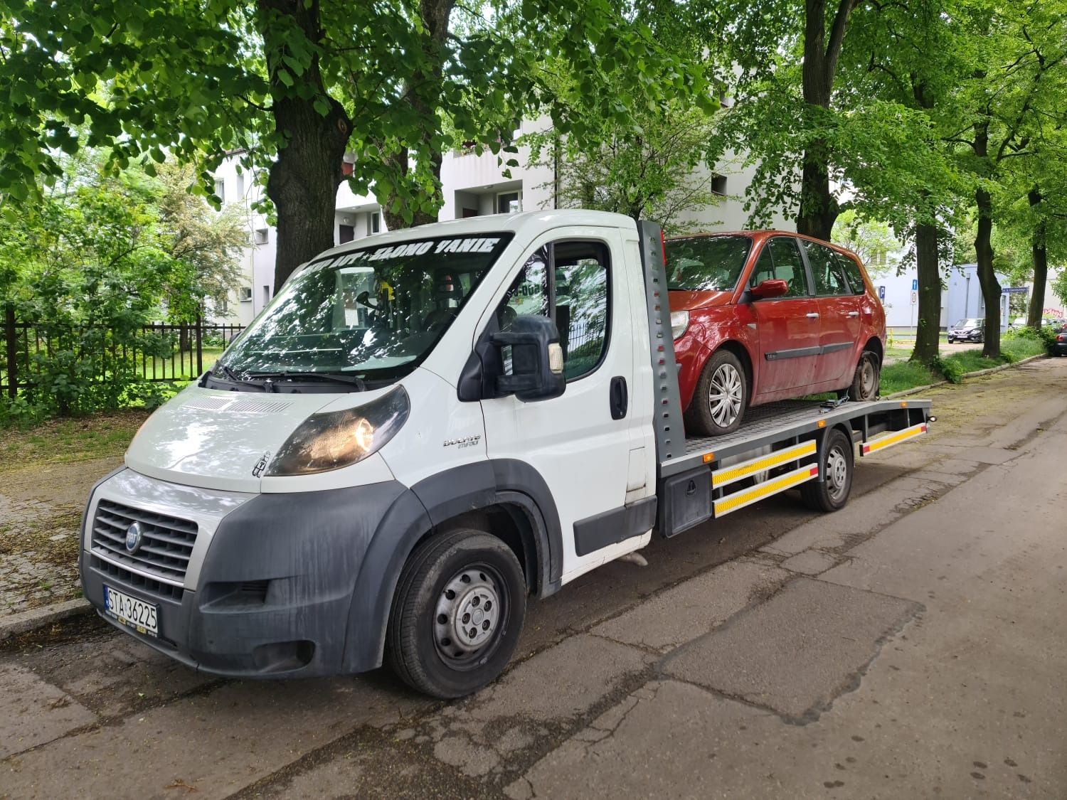 Biała laweta z czerwonym uszkodzonym samochodem osobowym na platformie, zaparkowana na ulicy w słoneczny dzień, widoczne drzewa i budynek w tle.