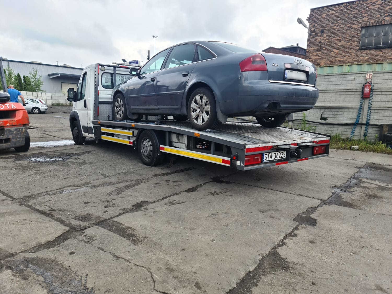 Szary samochód osobowy marki Audi A6 transportowany na lawecie, widoczny numer rejestracyjny pojazdu, w tle budynek z cegły i wózek widłowy.
