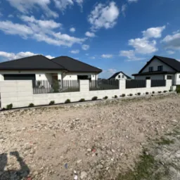 Nowoczesne ogrodzenie z metalowymi przęsłami i betonowymi słupkami, otaczające domy jednorodzinne w słoneczny dzień.