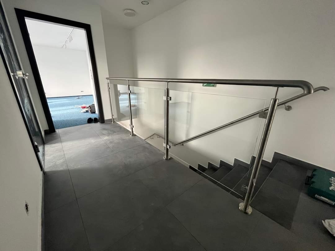 Wewnętrzne schody z nowoczesną balustradą ze stali nierdzewnej i szkła, widok z góry schodów na dolny poziom, z widocznym sprzętem montażowym i otwartymi drzwiami do pomieszczenia z niebieską...