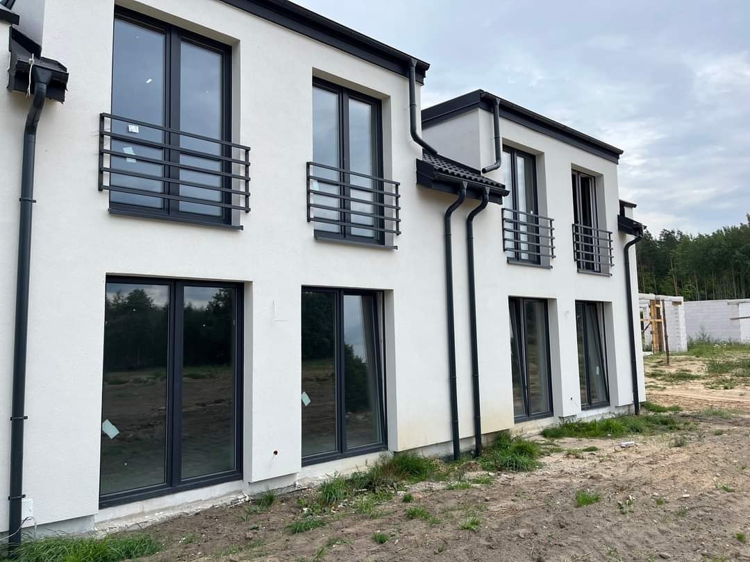 Elewacja nowo wybudowanego, dwukondygnacyjnego domu z czarnymi oknami i minimalistycznymi, poziomymi barierkami balkonowymi z metalowych profili.