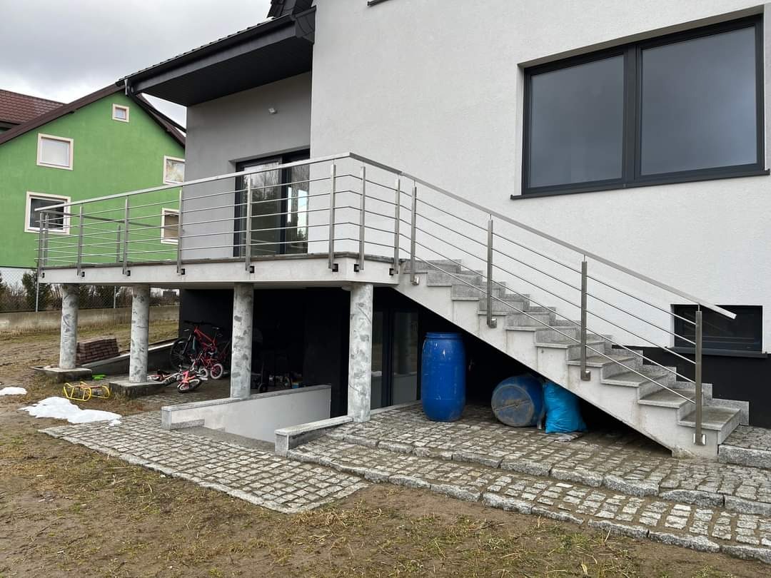 Zewnętrzne schody z balustradą nierdzewną w Ostródzie, prowadzące na taras nad garażem, wykończone kostką brukową.